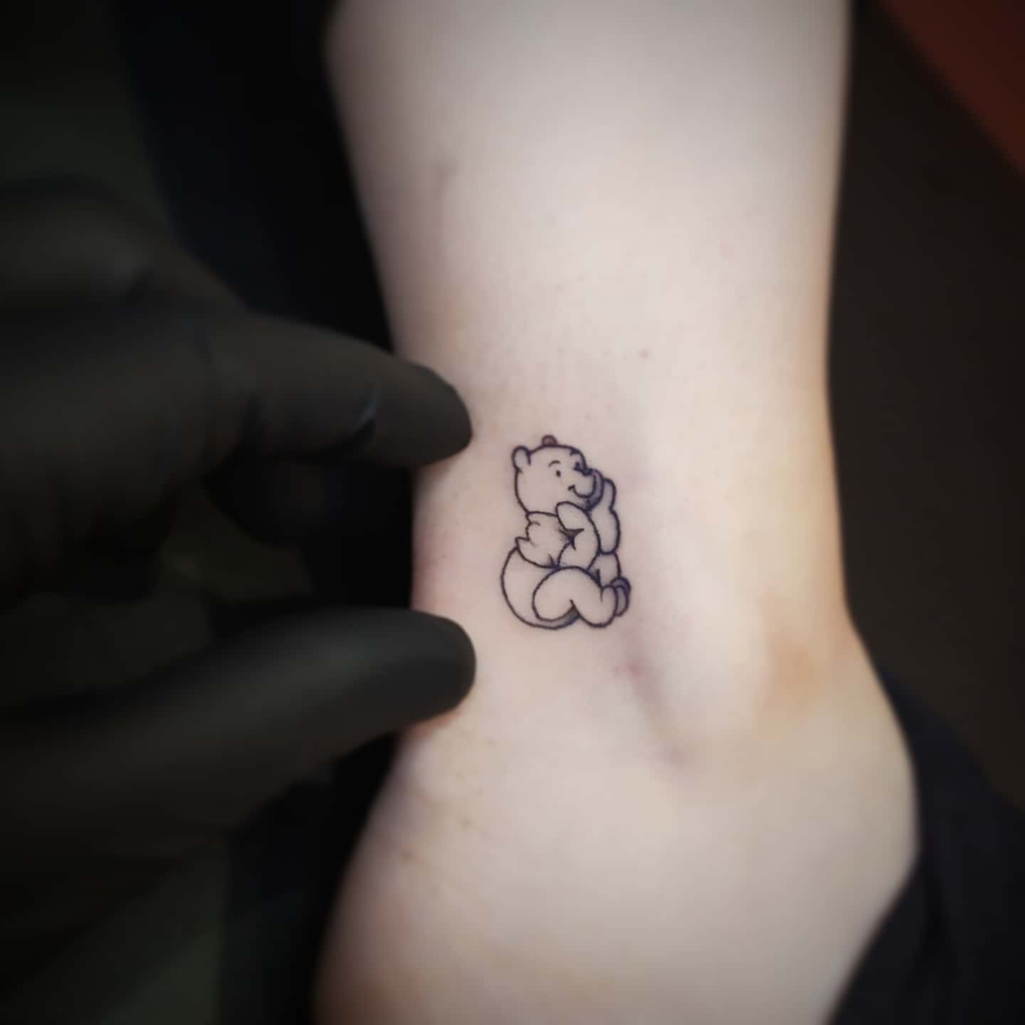 Tiny outline tattoo of Winnie the Pooh sitting with a thoughtful expression on a person's inner arm